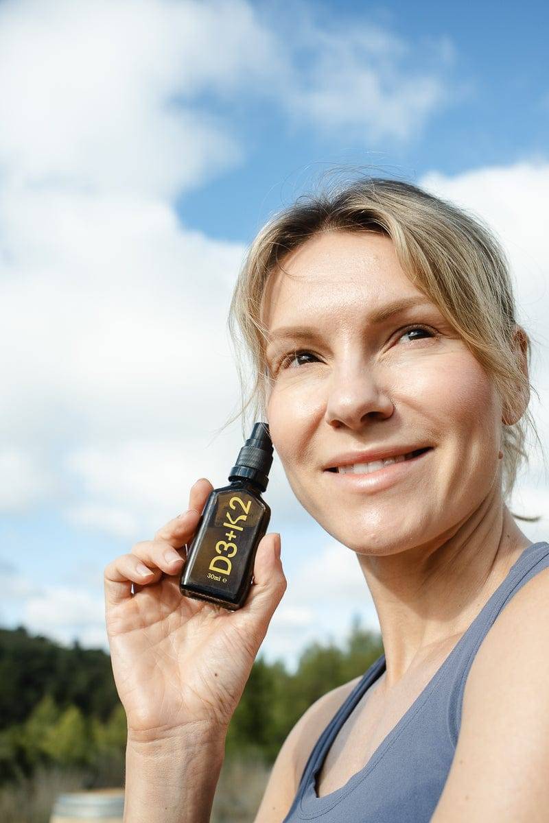 Smiling woman outdoors holding KURK Oral Spray - Vitamin D3 K2, promoting wellness and bone health with natural sunlight exposure.