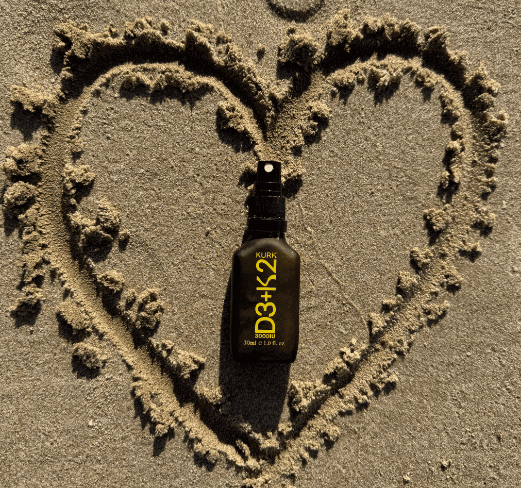 KURK Oral Spray - Vitamin D3 K2 bottle placed in a heart shape drawn in sand, promoting wellness and bone health with natural sunlight vibes.