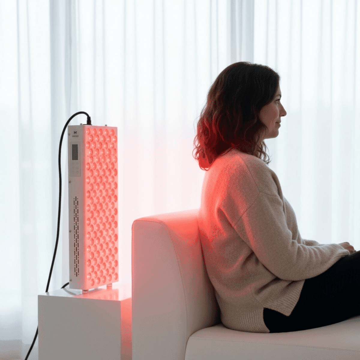 Woman seated beside Elite Series 506 Red Light Therapy Lamp, receiving back therapy for improved circulation and skin health in a bright room.